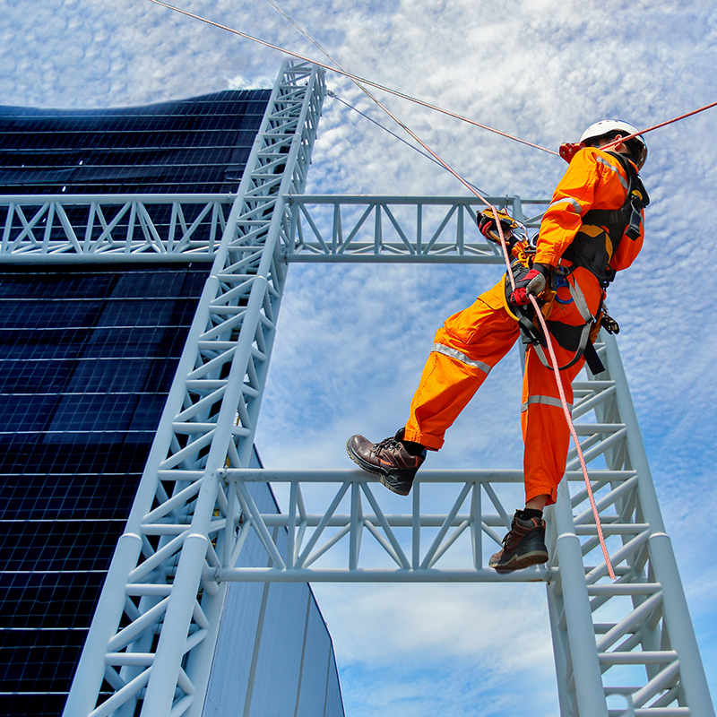 Arbeider i oransje vernedrakt og sele klatrer på hvit stålramme ved solcellepanel, blå himmel.
