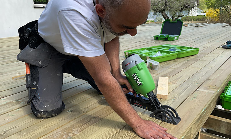 Håndverker på kne fester terrassebord med grønn ESSVE-spikerpistol ved åpen verktøykasse