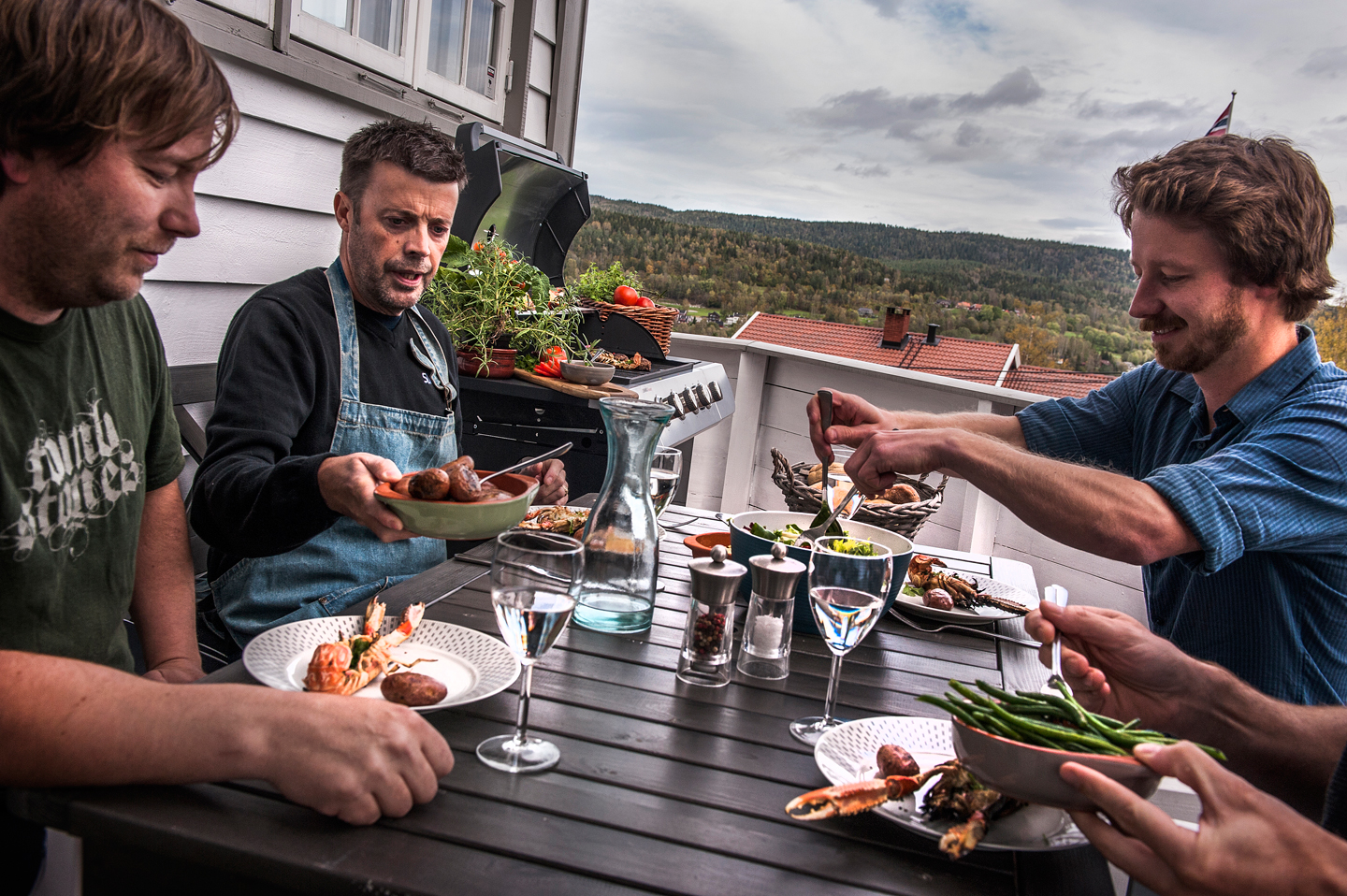 Personer rundt mørkt bord på hvit terrasse med sjømat, grønne bønner, poteter og vannkaraffel