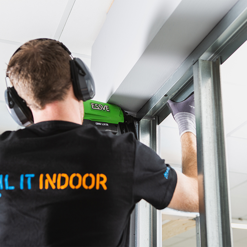 Arbeider med grønn Essve-spikerpistol mot stålprofil innendørs, iført hørselsvern.