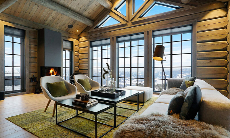 Lys tømmerstue med grå sofa, hvite lenestoler, grønt teppe, peis og fjellutsikt.