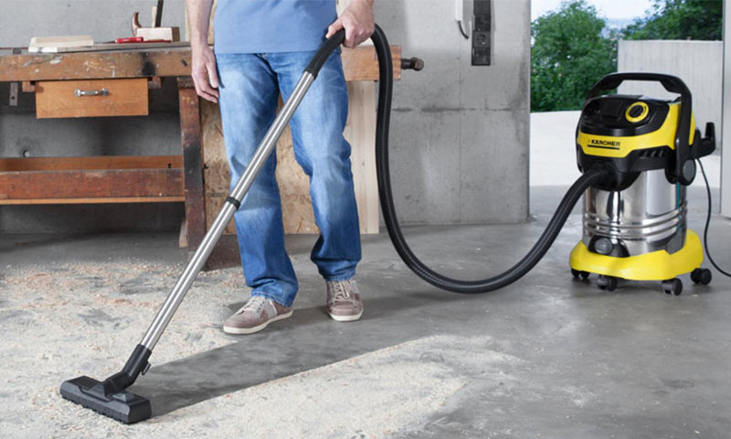 Mann i jeans bruker gul Karcher våt-/tørrstøvsuger på støvete betonggulv i verksted.