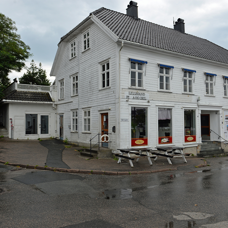 Hvit trebygning med blå markiser, utemøbler ved våt asfalt og skilt “Lillesand Pizza og Grill”.
