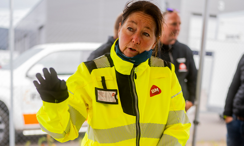 Person i gul refleksjakke med BM-logo vinker, kjøretøy og bygg i bakgrunnen.