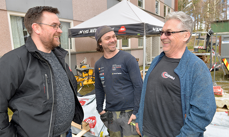Tre håndverkere i arbeidstøy ved husfasade, verktøy og BM-telt i bakgrunnen.
