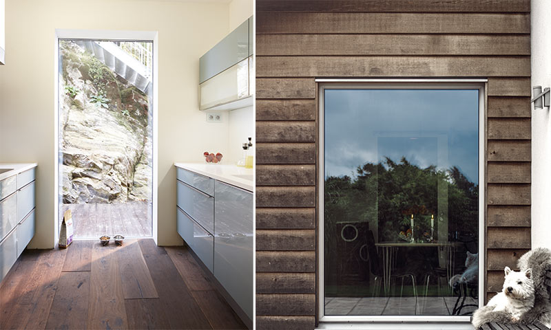 Lyst kjøkken med grå skap og dør ut mot stein; brun utvendig trevegg med vindu og hvit terrier