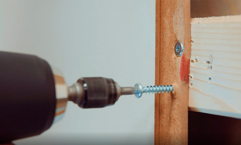 Nærbilde av svart drill som skrur sølvskrue inn i lyst treverk med rød malingsflekk mot hvit vegg