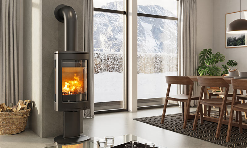 Svart vedovn med flammer ved vindu med utsikt til snødekt fjell, trebord og stoler.