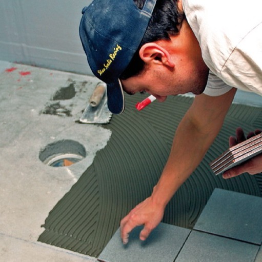 Håndverker med marineblå caps legger grå gulvfliser i mørk mørtel på betonggulv.