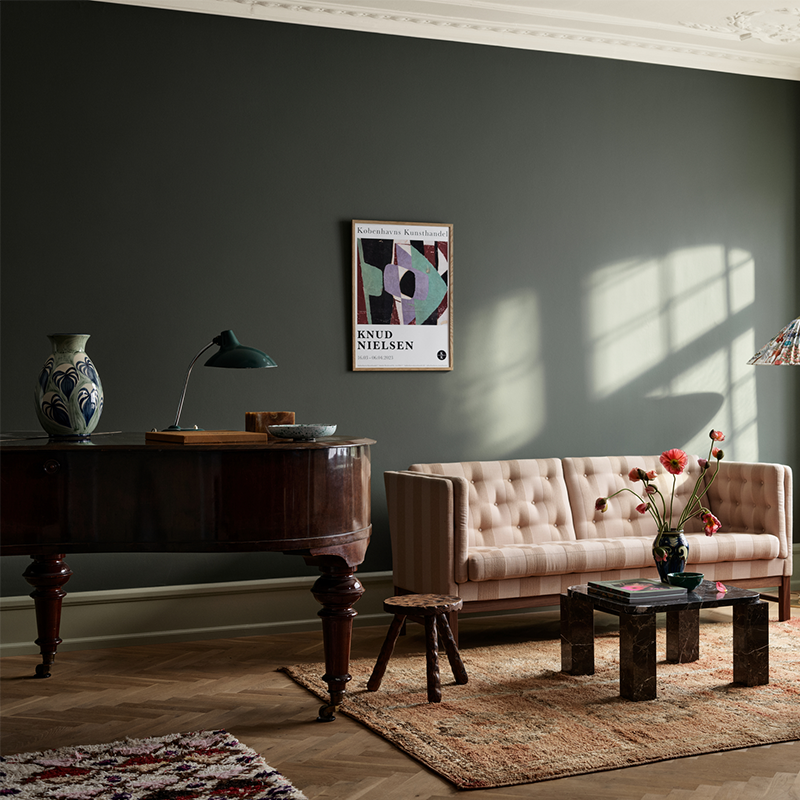 Elegant stue med mørkegrønne vegger, beige sofa, mørkt trebord, antikk skrivebord og kunstplakat i naturlig sollys.