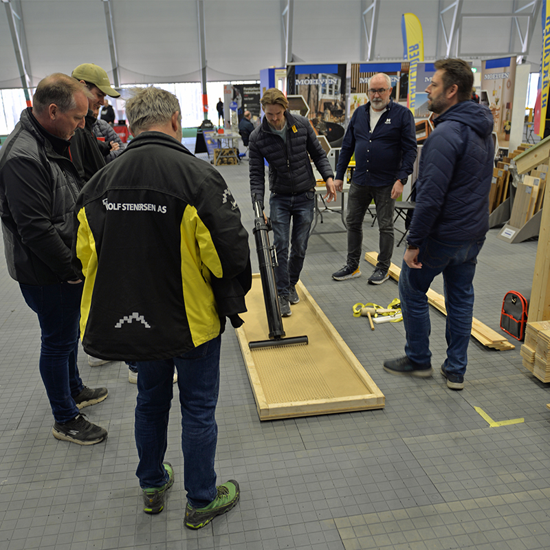 Demonstrasjon av verktøy på treplate i messehall, menn i blå og svart-gule jakker.