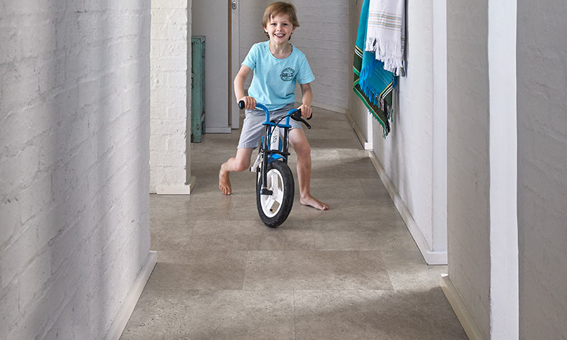 Barn kjører blå balansesykkel barføtt gjennom lys gang med hvit murvegg og hengt tøy.