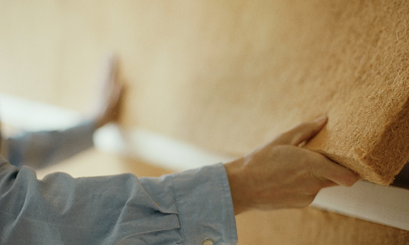 Hender i blå skjorte fester brun isolasjonsmatte på hvit veggskinne