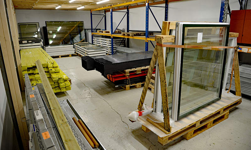 Lagerinteriør med glassvinduer på pall, gule trebjelker, blå pallereoler, metallplater.