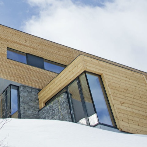 Moderne hytte i tre og stein med store vinduer, snø i forgrunnen og blå himmel