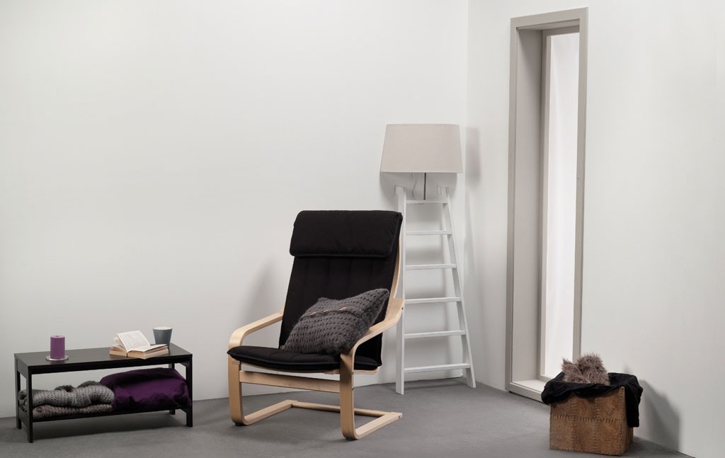 Minimalistisk hjørne med svart lenestol, grå pute, hvitt stigestativ-lampe, sidebord med lilla pledd.