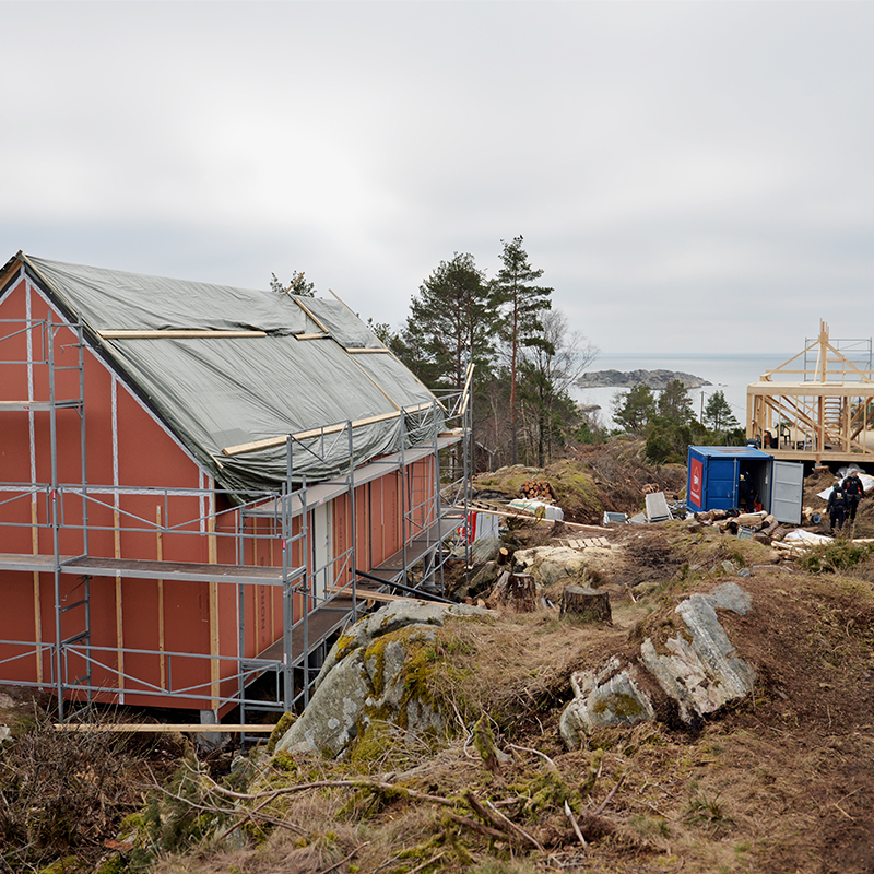 Rosa hus med stillas og trekonstruksjon på kystklippe med grantrær og grått hav