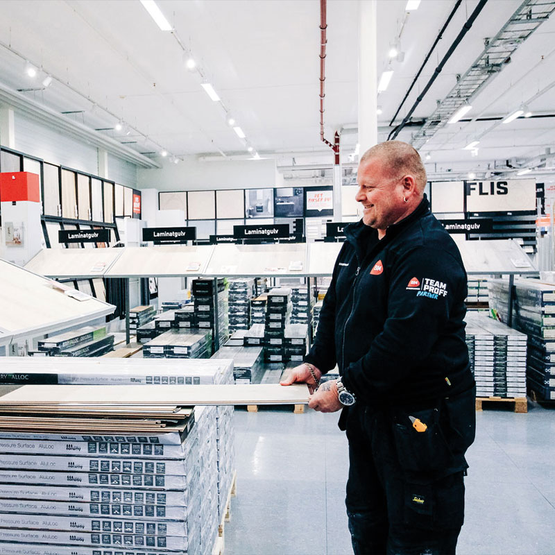 Ansatt i svart uniform holder laminatplanke foran hyller med laminatgulv i lys byggevarebutikk