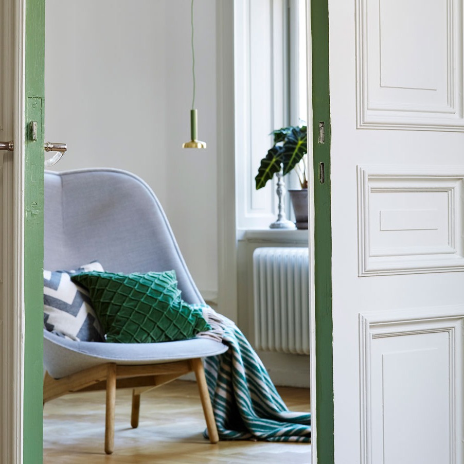 Åpen dør inn til lyst rom med grå lenestol, grønne puter, stripete pledd, hengelampe og plante