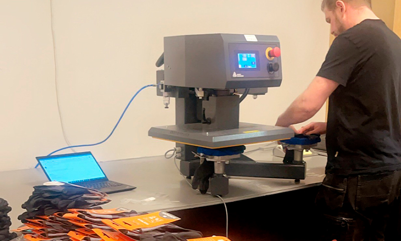 Svartkledd person bruker grå varmepresse på metallbord med laptop og tekstiler.