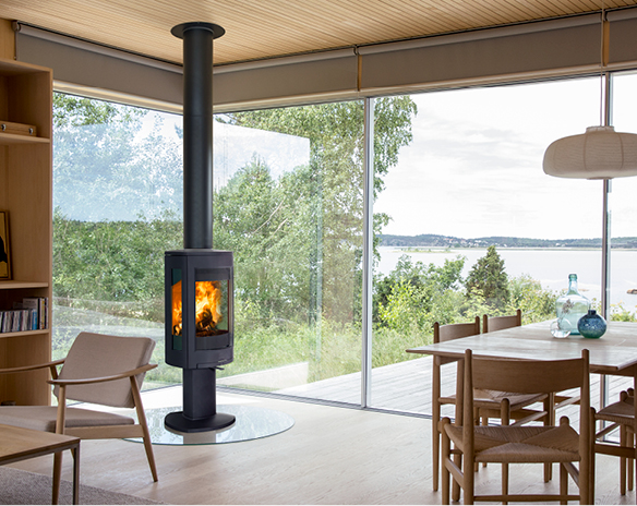 Lyst treinteriør med svart vedovn foran store vinduer mot grønn skog og innsjø.