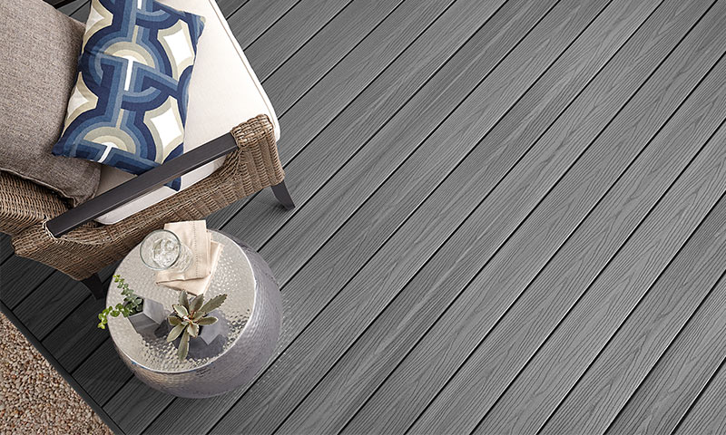 Ovenfra: grå terrassegulv, rottingstol med blå-hvit pute og sølvbord med sukkulent og glass.