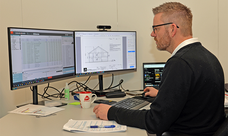 Mann i kontor med tre skjermer, hus-tegning på midtskjerm, rød kopp på pult.