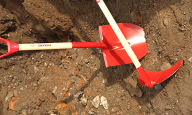 To røde hageredskaper (spade og hakke) med lyse treskaft på grusete jord.