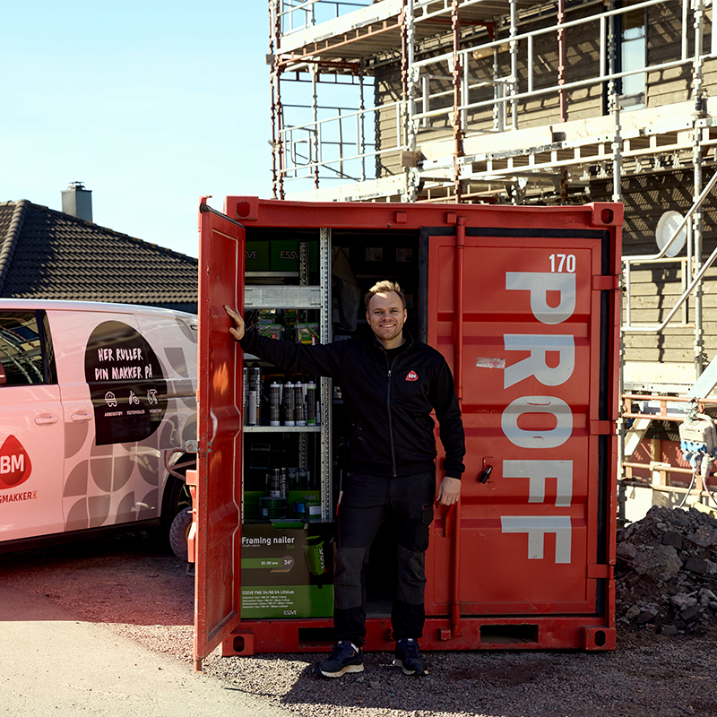Håndverker ved åpen rød container merket PROFF foran rosa-hvit varebil og stillas.