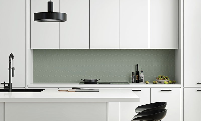 Moderne hvitt kjøkken med grønn heksagonal flis, sort pendellampe, svart kran og fruktskål.