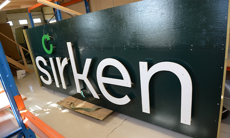 Hvit 3D-bokstavskilt 'sirken' med grønn pil på mørkegrønn planke i verkstedlager