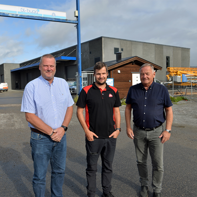 Tre menn i arbeidsklær står foran en grå lagerbygning med blått skilt, liten hytte og gul kran.