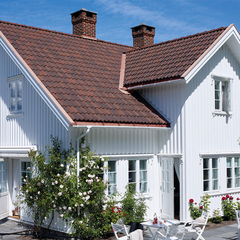 Hvitt trehus med brunt tegltak, blomstrende rosebusker og lite hagebord med stoler.