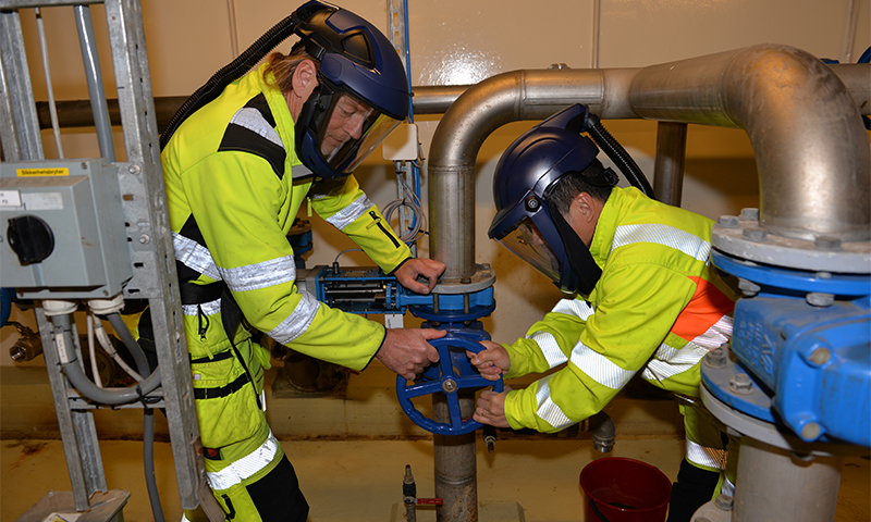 To arbeidere i gule vernedrakter åpner blå ventil på metallrør i industrilokale