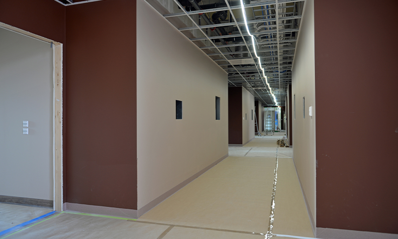 Lang korridor under oppussing med beige gulv, brune vegger, eksponert tak og lysrør