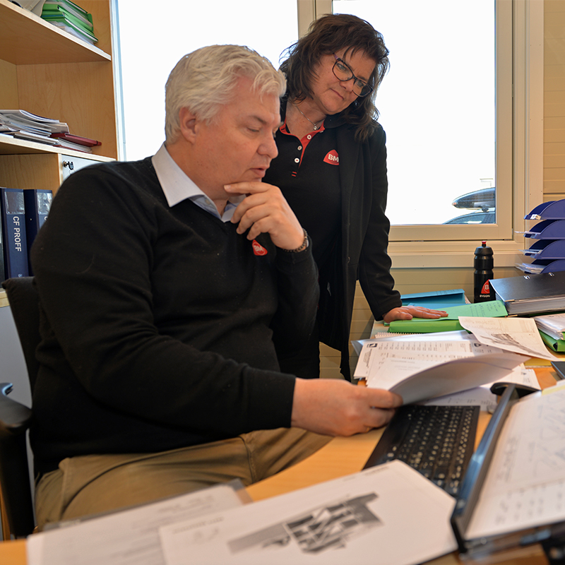 Mann og kvinne ved kontorpult med papirer, datatastatur, grønn mappe og vindu.