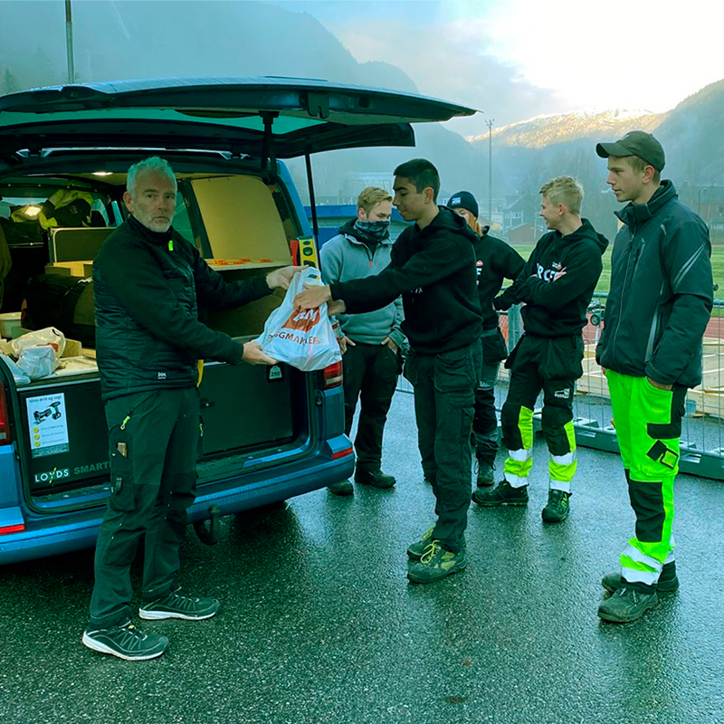 Mann ved åpen blå varebil gir pose til arbeidsgruppe i svart og neongrønt tøy foran fjell.