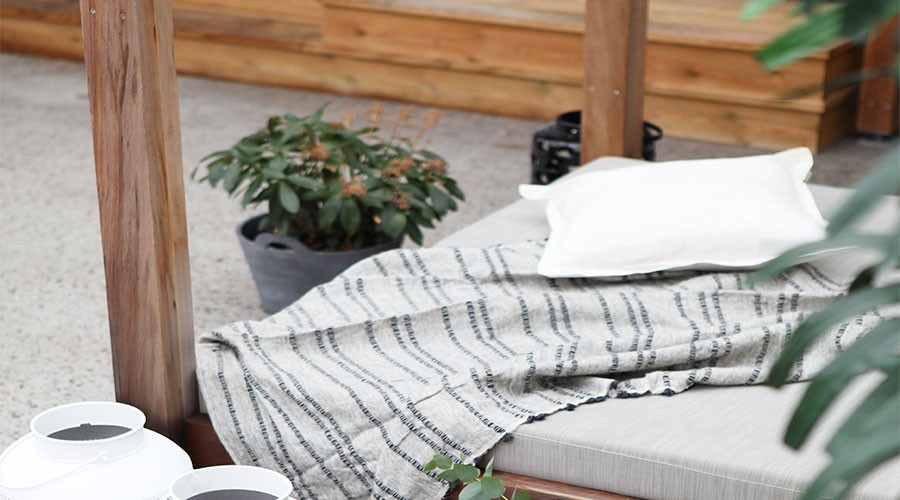 Utendørs loungebed i lyst tre med grå madrass, stripete pledd, hvit pute og grønne planter.