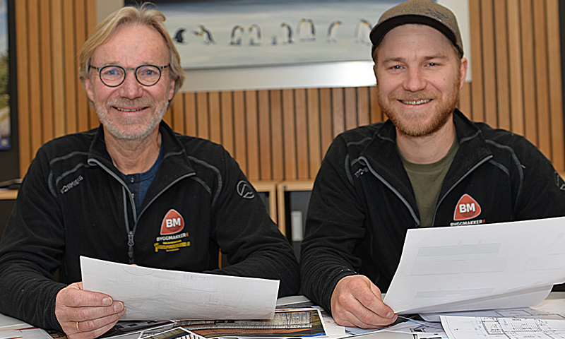 To menn i svarte BM-jakker sitter ved bord med byggeplaner, trepanelvegger og pingvinbilde bak.