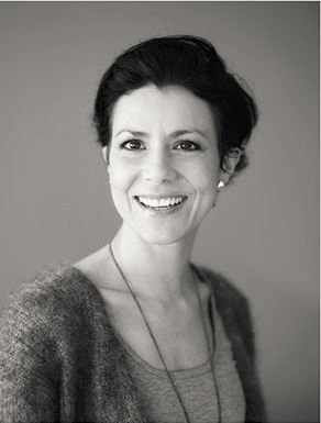 Svart-hvitt portrett av person med kort hår, strikket cardigan, singlet og ørepropper.