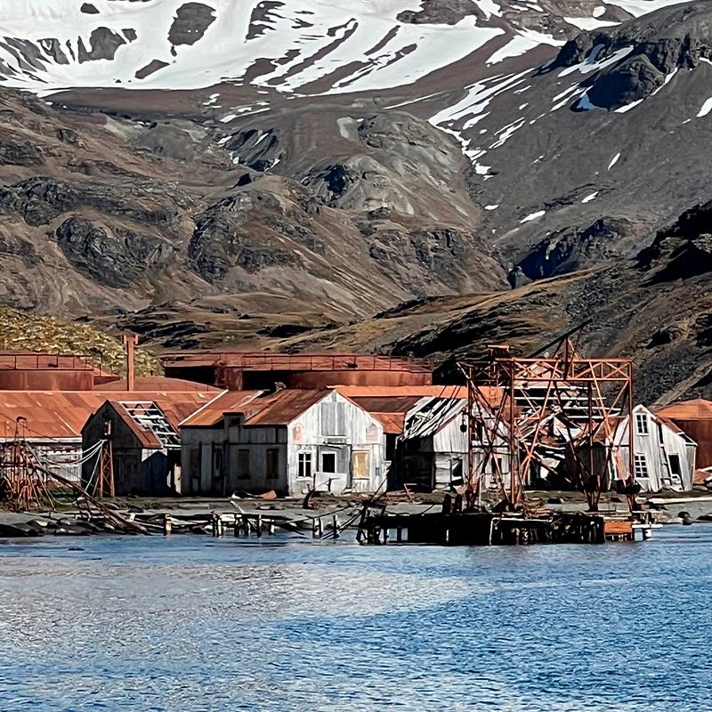 Forlatte rustne fabrikksbygninger ved blått vann med snødekte brune fjellsider i bakgrunnen.