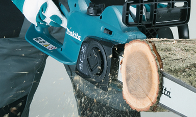 Hender med hvite og grønne hansker holder turkis Makita-kjedesag som sager i tre, flis spruter