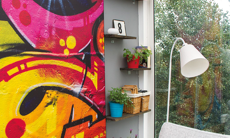 Interiørhjørne: rosa-gul grafittivegg, hyller med planter og kurver, vindu mot hage, hvit gulvlampe