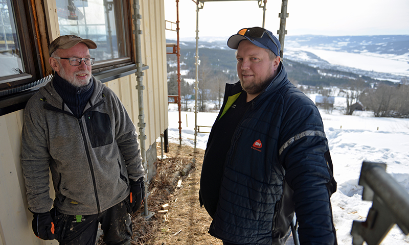 To håndverkere i grå og blå jakker på stillas ved et hus med snødekt dal i bakgrunnen.