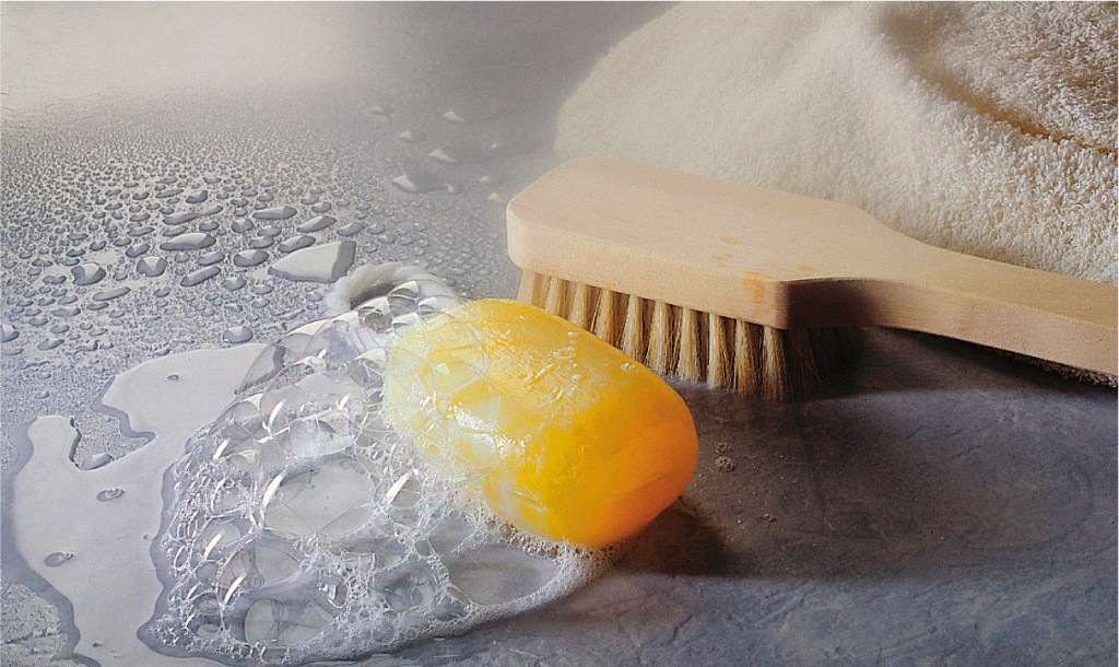 Gul såpestykke med hvitt skum ved siden av en treskrubbebørste på grå, våt overflate.