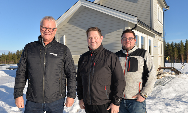 Tre menn i vinterjakker foran nybygd grå trevilla, snø og furuskog under blå himmel