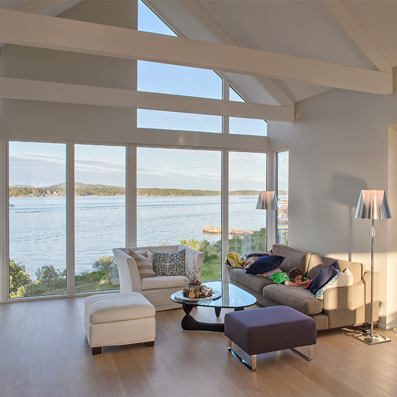 Lyst stueinteriør med store vinduer mot innsjø, sofa med sovende barn og hvit lenestol.