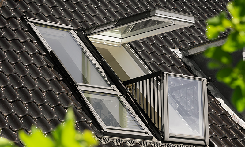 Sort glinsende tak med to hvite, åpne takvinduer, grønt løv i forgrunnen.