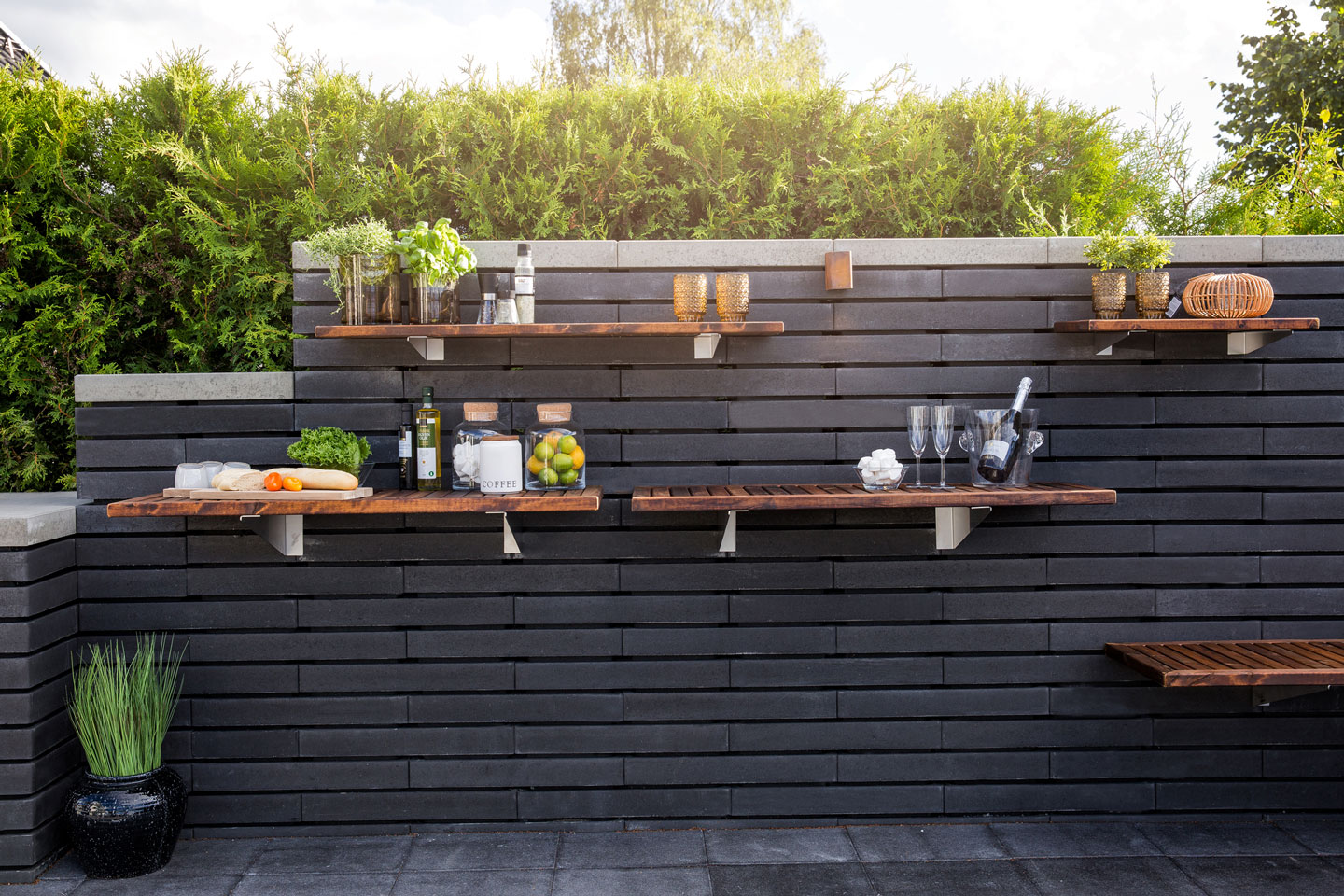 Mørk murvegg med flytende trehyller pyntet med urter, frukt, kjøkkenutstyr og champagneglass.