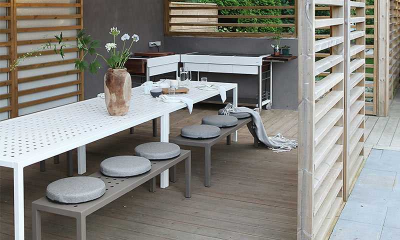 Minimalistisk uteplass: hvitt perforert bord, grå benker med runde puter, treespalier.
